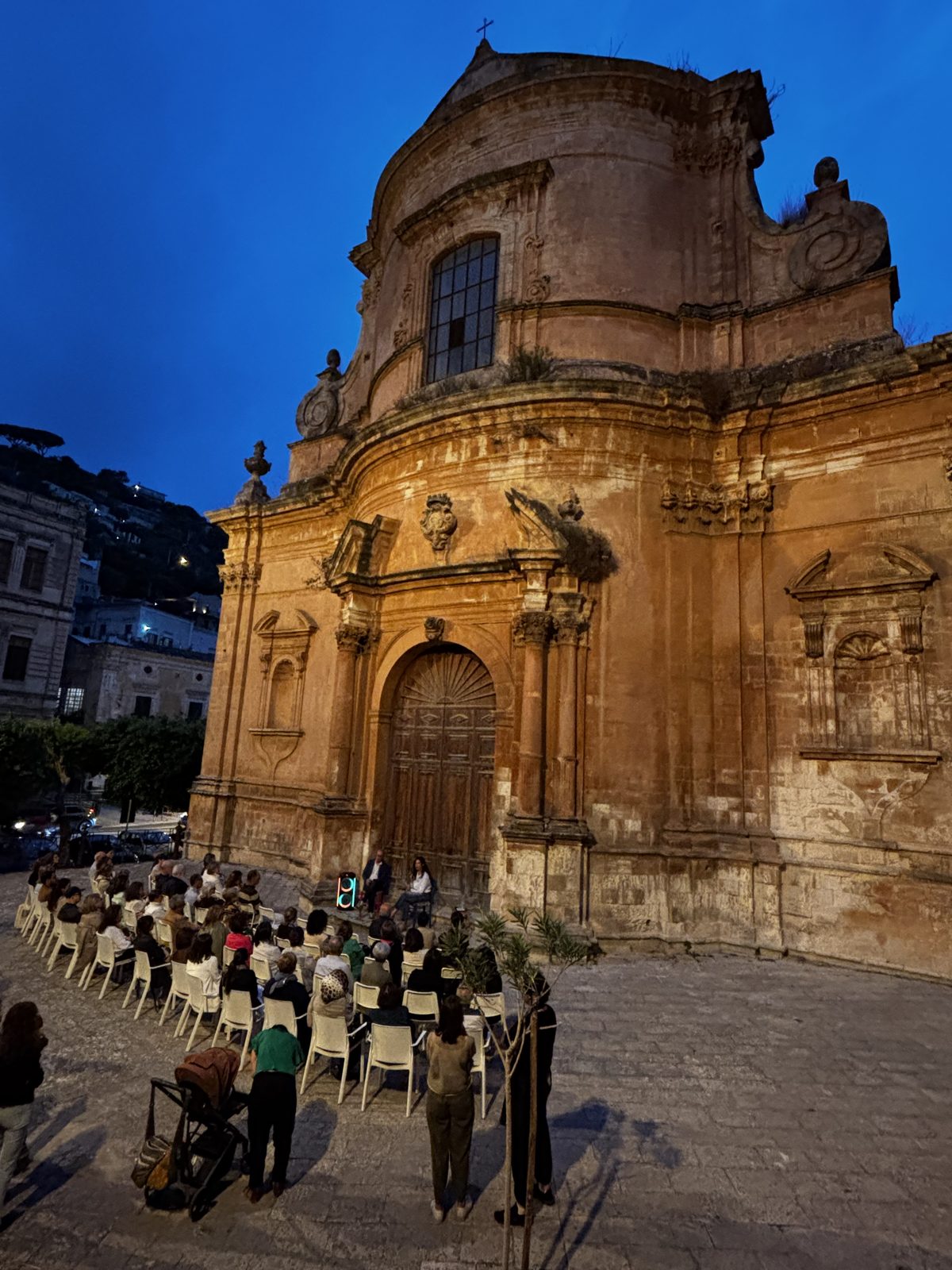 Il pubblico seduto davanti a una chiesa barocca all’imbrunire, durante un incontro all’aperto