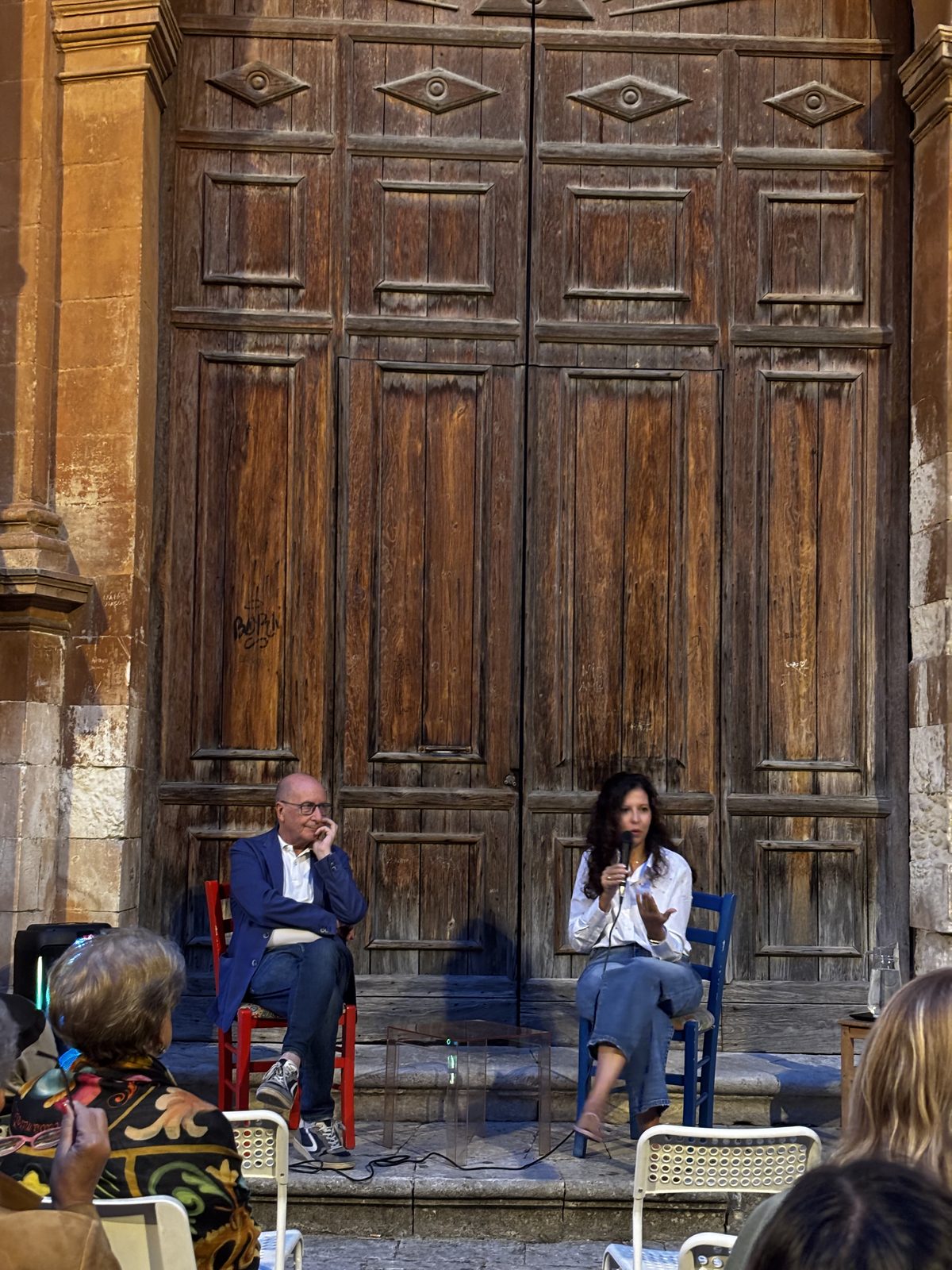 Miryam Cannizzaro e un altro relatore seduti davanti a un portone di legno durante un incontro pubblico